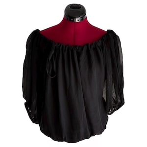 May include: Black off-the-shoulder blouse with long, sheer sleeves. The top is made of a lightweight, flowing fabric and features a gathered neckline and elastic cuffs. The blouse is displayed on a red dress form.