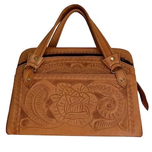 May include: A tan leather handbag with a floral design. The bag features a large embossed rose in the center, surrounded by intricate patterns. It has two top handles and a zipper closure. The bag is rectangular and has a small side strap.