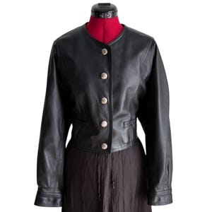 May include: A black leather jacket featuring a round neckline, long sleeves, and a front button closure with five silver-toned metallic buttons. The jacket has a cropped silhouette with horizontal seam detailing at the waist and two small pockets.