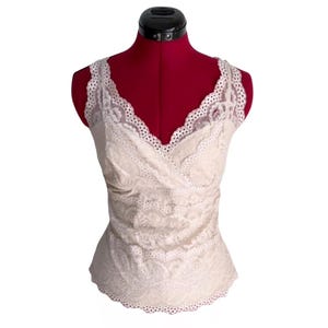 May include: Cream-colored lace camisole with a V-neckline and scalloped edges. The top is displayed on a red dress form. The delicate lace pattern covers the entire garment, creating a romantic and feminine look. The camisole is sleeveless.
