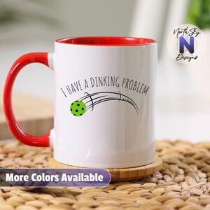 May include: White ceramic mug with a red interior and handle. The mug features the text "I HAVE A DINKING PROBLEM" with a graphic of a green pickleball. The words "More Colors Available" are at the bottom.