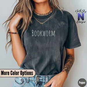 May include: A dark gray t-shirt with the word "BOOKWORM" printed in white on the front. The shirt is being worn by a person with a tattoo on their arm.