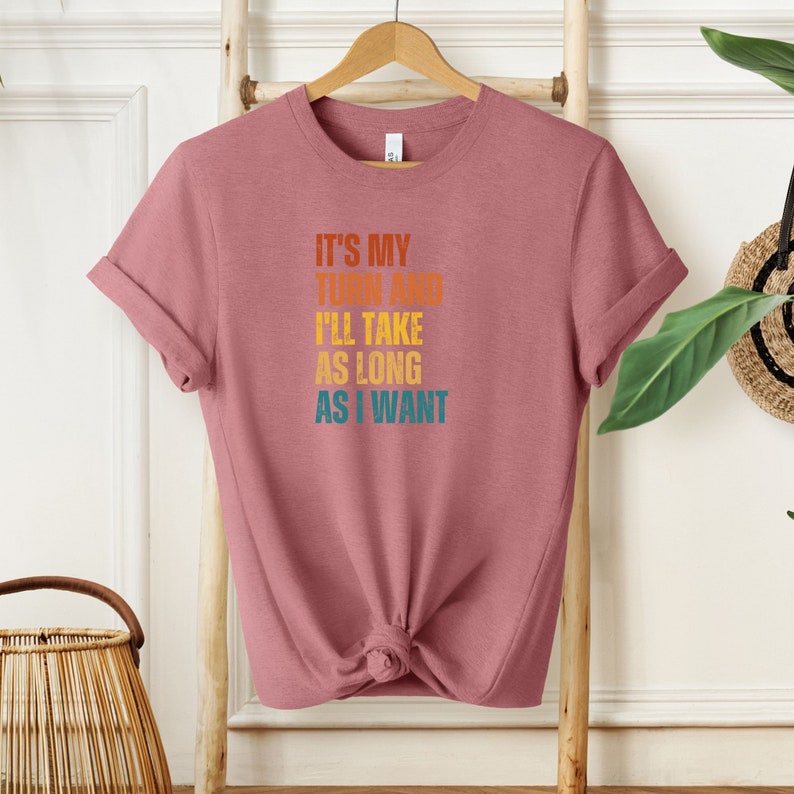 K&ouml;nnte beinhalten: Ein pinkfarbenes T-Shirt mit dem Text "It's my turn and I'll take as long as I want" in einer Retro-Schriftart mit orange, gelben und blauen Farben.