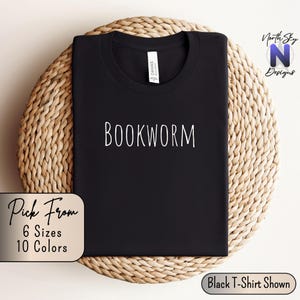 May include: Black t-shirt with the text "BOOKWORM" in white lettering. The t-shirt is displayed on a woven surface. The text "Pick From 6 Sizes 10 Colors" is visible in the lower left corner of the image. The text "Black T-Shirt Shown" is visible in the lower right corner of the image.