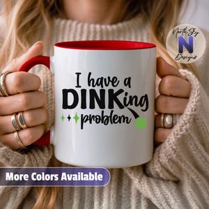 May include: White ceramic mug with a red interior and handle. The mug features the text "I have a DINKing problem" in black, with green accents and a green pickleball graphic. The words "More Colors Available" are at the bottom.