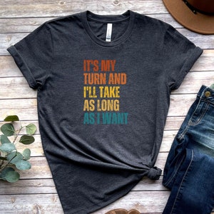 K&ouml;nnte beinhalten: Ein dunkelgraues T-Shirt mit einer Grafik im Vintage-Stil, auf dem "It's my turn and I'll take as long as I want" steht.