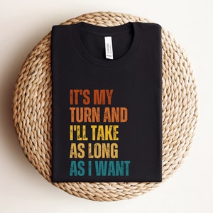 May include: Black t-shirt with a vintage-style graphic that reads "It's my turn and I'll take as long as I want."
