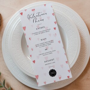 Może przedstawiać: Menu walentynkowe z wzorem różowych i białych serc. Menu zawiera przystawki, dania główne i deser. Tekst brzmi: "Menu walentynkowe", "Przystawka", "Zupa z dyni podawana z kromką czosnkowego chleba", "Danie główne", "Wolno gotowana wołowina z gotowanymi na parze warzywami i frytkami", "lub", "Kotlet z kurczaka z sałatką i frytkami", "Deser", "Tort urodzinowy".