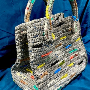 May include: A gray crocheted tote bag with colorful accents. The bag has two handles and a rectangular shape.