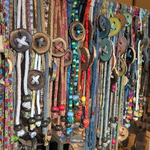 May include: A display of handmade necklaces with wooden button pendants and colorful beaded accents. The necklaces feature various cord colors, including white, red, blue, and green. The buttons have cross-shaped designs.