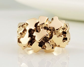 May include: A gold ring with a textured, nugget-like design. The ring features a wide band and a surface made up of irregular, overlapping shapes. The ring is shown on a white surface, with soft lighting enhancing its metallic sheen.