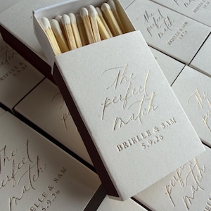 May include: A rectangular matchbox with a cream-coloured cover featuring gold foil text that reads The perfect match, BRIELLE & SAM, and 5.9.26. The box is partially open, revealing wooden matches with white tips, surrounded by identical matchboxes.