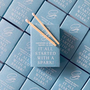 Peut inclure: Une collection de boîtes d'allumettes bleu clair disposées en grille, chacune portant le texte "IT ALL STARTED WITH A SPARK!", les noms "CRYSTEN & BRETT", la date "September, 24, 2025" et un monogramme. Deux allumettes en bois reposent sur la boîte centrale.