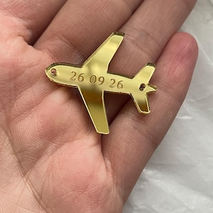 May include: A gold-coloured airplane charm with the engraved date "26 09 26". The charm has a polished finish and is held in a hand. The airplane has a classic design with wings and a tail.