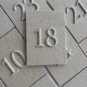 Pode incluir: Uma coleção de azulejos retangulares bege texturizados com números brancos em relevo. O azulejo central exibe o número 18. Outros azulejos mostram números como 10, 21 e 23. Os azulejos estão dispostos sobre uma superfície branca.