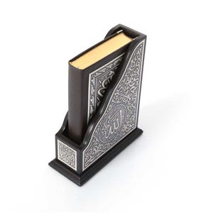 May include: A dark brown wooden book holder with ornate silver detailing. The holder cradles a book with gold-edged pages. The silver design features Arabic calligraphy and floral patterns, set against a white background.