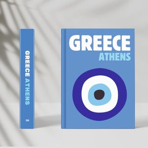 Könnte beinhalten: Ein blaues Buch mit den Worten "GREECE ATHENS" in Weiß. Das Cover zeigt ein großes blaues und weißes Augensymbol. Der Buchrücken zeigt ebenfalls den Text "GREECE ATHENS".