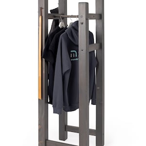 Könnte beinhalten: Ein dunkelgrauer Holz-Kleiderständer mit zwei horizontalen Stangen und einem Hutständer oben. Der Ständer hält einen schwarzen Hoodie mit einem blauen Logo, auf dem "m wooden" steht, und ein schwarzes T-Shirt. Eine graue Baseballkappe mit dem Text "H2O" befindet sich auf dem Hutständer.