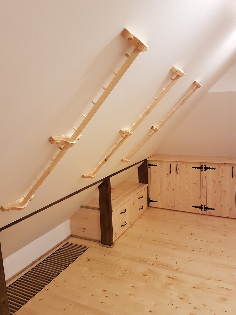 May include: Four wooden climbing bars mounted on a white wall in an attic room. The bars are different lengths and are made of natural wood. The room has a wooden floor and a wooden cabinet with doors.