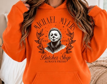 May include: A bright orange hooded sweatshirt featuring a black graphic print of Michael Myers. The design includes the text "MICHAEL MYERS", "EST. 1978", "BUTCHER SHOP", and "ALWAYS FRESH" surrounded by a leafy wreath and small stars.