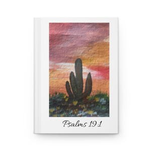 May include: A white hardcover journal featuring a rectangular painting of a dark green cactus silhouette against a vibrant sunset sky with shades of pink, orange, and yellow. The text Psalms 19:1 is printed in black in a script font below the image.