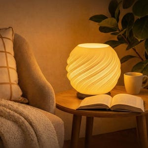 Könnte beinhalten: Eine kugelförmige Tischlampe mit einem strukturierten, wirbelnden Design strahlt ein warmes Licht aus. Sie steht auf einem kleinen Holztisch neben einem offenen Buch und einer gelben Tasse. Ein beiger Sessel und eine Decke sind im Hintergrund.