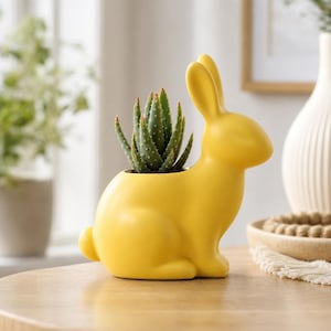 Op de afbeelding: Een felgele keramische plantenbak in de vorm van een zittend konijn. Het konijn heeft lange oren en een gladde, matte afwerking. Een kleine groene vetplant met puntige, getextureerde bladeren is geplant in de holle rug van het konijn.