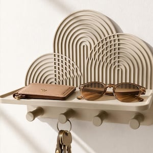 Op de afbeelding: Een beige wandplank met een decoratief achterpaneel met concentrische boogpatronen. De plank heeft vier cilindrische haken aan de onderkant, waaraan een sleutelbos hangt. Een kleine beige portemonnee en een bruine zonnebril liggen op het oppervlak.