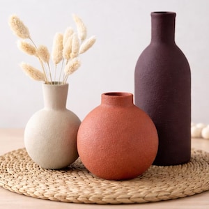 Könnte beinhalten: Drei strukturierte Vasen in Creme, Terrakotta und Dunkellila. Die cremefarbene Vase ist kugelförmig mit einem schmalen Hals und enthält getrocknete beige Grashalme. Die terrakottafarbene Vase ist rund mit kurzem Hals, die lilafarbene Vase ist hoch und zylindrisch.