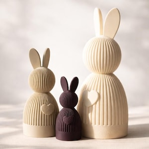 Könnte beinhalten: Drei Hasenfiguren mit gerippter Textur und abgerundeten Körpern. Zwei sind cremefarben und eine ist dunkelbraun. Jeder Hase hat ein kleines herzförmiges Detail auf der Brust und aufrecht stehende Ohren in verschiedenen Größen.
