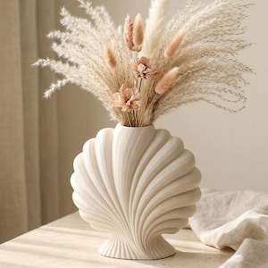 Könnte beinhalten: Eine matte, cremeweiße Keramikvase in Form einer großen Jakobsmuschel mit tiefen vertikalen Rillen. Die Vase enthält ein Trockenblumenarrangement mit flauschigem Pampasgras, kleinen rosa-beigen Hasenschwänzchen und zwei kleinen Trockenblumen.