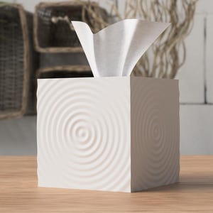 Peut inclure: Une boîte à mouchoirs carrée blanche ornée d'un motif en relief de cercles concentriques sur ses faces extérieures. Un mouchoir en papier blanc est tiré vers le haut depuis l'ouverture centrale, dépassant du bord supérieur de la boîte.