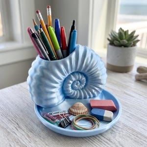 Könnte beinhalten: Ein hellblauer Schreibtisch-Organizer in Form eines Nautilus-Gehäuses mit einer runden Basis. Der Gehäuseteil enthält verschiedene Stifte, während auf der Ablage eine kleine Muschel, ein rosa Radiergummi, ein blau-weißer Radiergummi, Büroklammern und Gummibänder liegen.