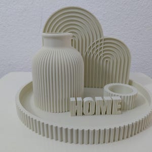 May include: A cream-coloured decorative set featuring a round ribbed tray, a ribbed vase, two arched ribbed panels, a small ribbed circular container, and a block sign with the word HOME in raised letters, all sharing a consistent ribbed texture.