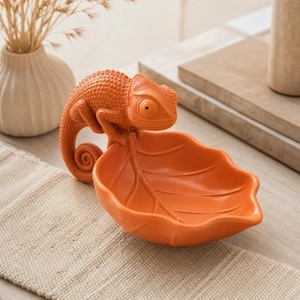 Könnte beinhalten: Eine leuchtend orangefarbene Keramikschale in Blattform mit einer texturierten Chamäleonfigur am Rand. Das Chamäleon hat einen geringelten Schwanz und eine detaillierte Hautstruktur, während die blattförmige Schale ausgeprägte Blattadern zeigt.