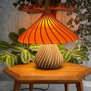 May include: A mushroom-shaped table lamp featuring a vibrant orange, spiraled, wide-brimmed shade and a textured, off-white, spiraled base. The lamp emits a warm, glowing light from beneath the shade, creating a distinct, layered, and ribbed appearance.