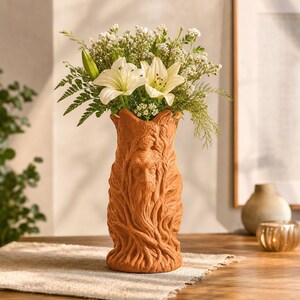 Könnte beinhalten: Eine terrakottafarbene Vase mit einem skulpturalen Relief eines bärtigen Gesichts, umgeben von detaillierten Baumrinden- und Blattstrukturen. Die Vase ist mit einem Strauß aus weißen Lilien, kleinen weißen Blüten und grünem Farn gefüllt.