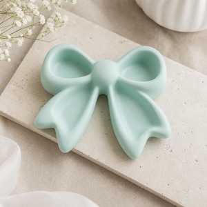 Peut inclure: Un plateau décoratif vert menthe clair en forme de nœud. Le plateau présente une finition lisse et mate avec un nœud central et deux boucles symétriques s'étendant vers l'extérieur, créant une surface concave peu profonde pour les petits objets.