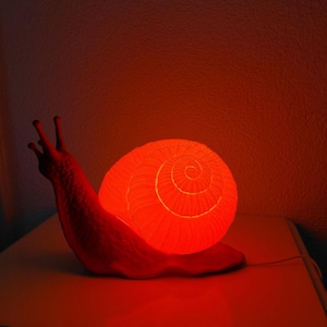 May include: A snail-shaped lamp emitting a vibrant red glow. The lamp features a textured, spiraled shell and a smooth body with two prominent eye stalks extending upward. A thin white power cord extends from the base of the snail figure.
