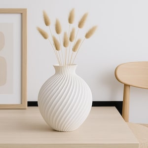 Könnte beinhalten: Eine weiße Keramikvase mit Spiralmuster hält getrocknete beige Blumen. Die Vase steht auf einem hellen Holztisch, mit einem gerahmten abstrakten Kunstdruck und einer kleinen weißen Tasse in der Nähe. Im Hintergrund befindet sich ein Holzstuhl.