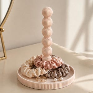 Pode incluir: Um suporte de joias rosa pálido com um pilar vertical canelado sobre uma base circular. O suporte contém vários elásticos de cabelo de tecido em tons de creme, rosa antigo e taupe, além de um pequeno anel dourado e brincos de pérola.