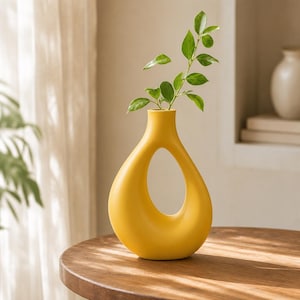 Peut inclure: Un vase en céramique jaune vif avec une finition mate lisse et une forme de goutte d'eau distinctive comportant une grande découpe ovale creuse au centre. Une branche feuillue verte est placée dans le col étroit du vase.