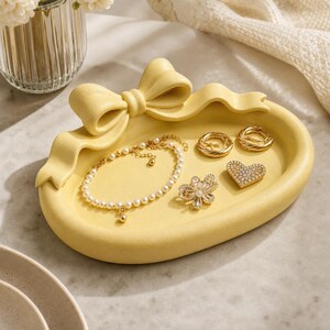 May include: A pale yellow oval jewelry tray featuring a sculpted bow detail on the top edge. The tray holds a white pearl bracelet, two gold-toned hoop rings, a gold-toned flower-shaped brooch with clear crystals, and a heart-shaped pin.