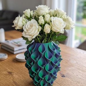 Könnte beinhalten: Eine dekorative Vase in Teal- und Lilatönen mit einem geschichteten Design, die einen Strauß weißer Rosen enthält. Die Vase steht auf einem Holztisch und präsentiert ein modernes Blumenarrangement.