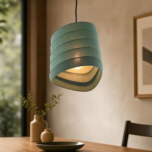 Op de afbeelding: Een blauwgroene hanglamp met een getextureerde, geribbelde en licht asymmetrische cilindrische kap. De lamp hangt aan een zwart snoer en verspreidt een warme, zachte gloed vanuit de binnenkant, die is voorzien van een gladde, lichtgekleurde voering.