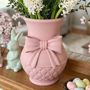 Könnte beinhalten: Eine matte, pastellrosa Keramikvase mit einer markanten, skulpturalen Schleife auf der Vorderseite und einem strukturierten Steppmuster am Fuß. Die Vase hat einen weit ausgestellten Rand und einen bauchigen Körper für Blumenarrangements.