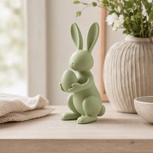 Könnte beinhalten: Eine matte, salbeigrüne Hasenfigur, die aufrecht auf einer flachen Oberfläche steht. Der Hase hat lange, spitze Ohren und hält ein glattes, eiförmiges Objekt in seinen Vorderpfoten. Die Skulptur hat eine einheitliche Farbe und Oberfläche.
