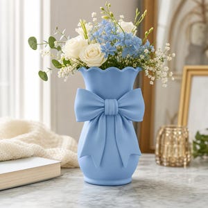 Könnte beinhalten: Eine hellblaue Vase mit gewelltem Rand und einer großen, auffälligen Schleife auf der Vorderseite. Die Vase ist mit einem Strauß aus weißen Rosen, hellblauen Ritterspornblüten sowie kleinen Zweigen aus Eukalyptus und Schleierkraut gefüllt.