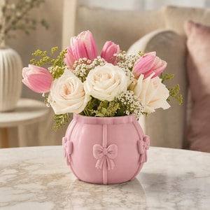 Könnte beinhalten: Eine runde, hellrosa Keramikvase mit vertikalen Rillen und dreidimensionalen Schleifenverzierungen. Die Vase ist mit einem Blumenarrangement aus zartrosa Tulpen, cremeweißen Rosen und kleinen weißen Schleierkrautblüten gefüllt.