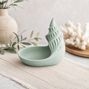 Könnte beinhalten: Eine matte, salbeigrüne Keramikschale in Form einer Muschel. Die Schale verfügt über ein glattes, abgerundetes Becken zur Aufbewahrung kleiner Gegenstände und eine strukturierte, spiralförmige Muschelstruktur an einer Seite in einem sanften Grünton.